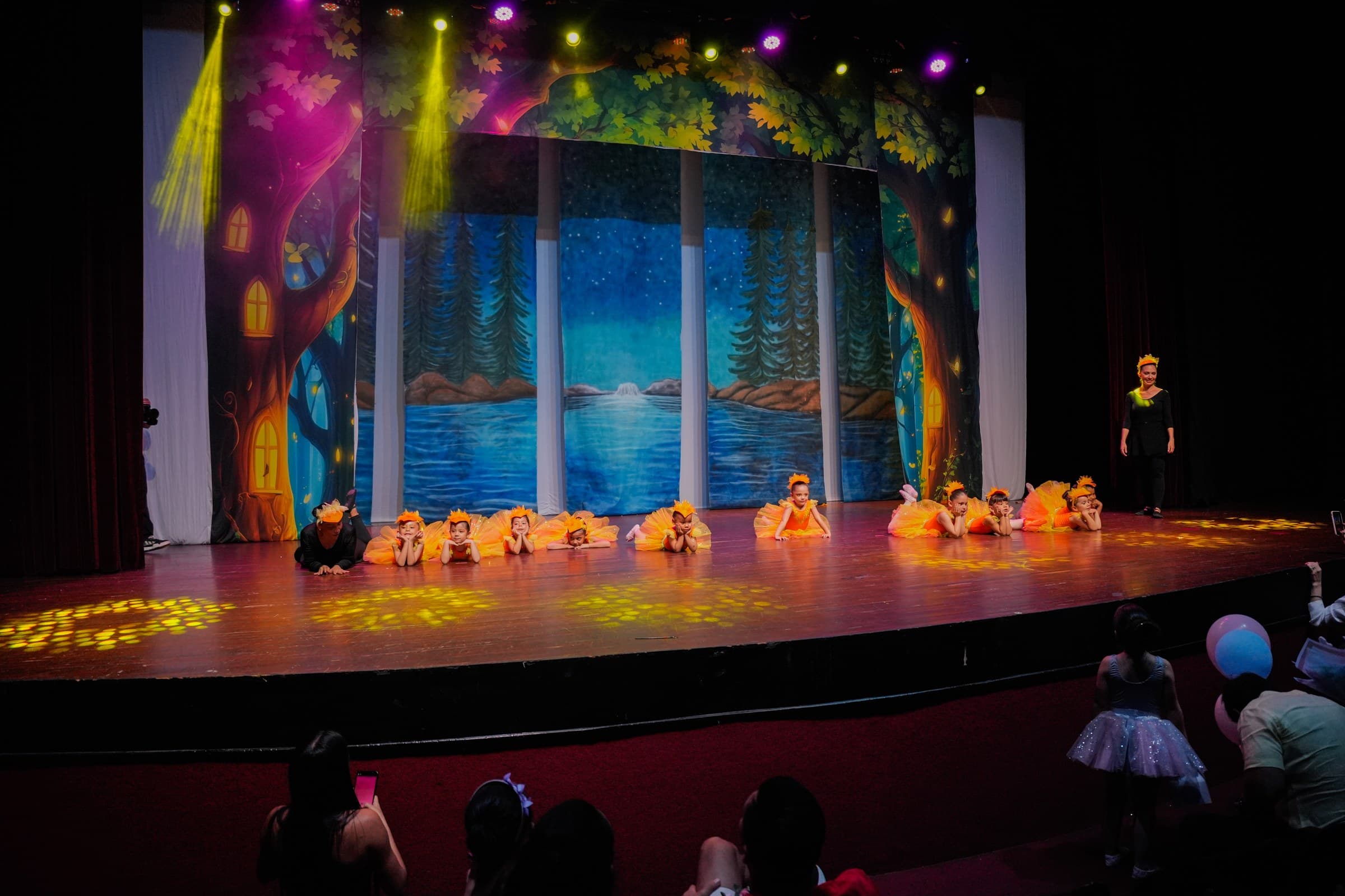 Niñas bailarinas con vestuario naranja frente a un decorado de bosque y lago