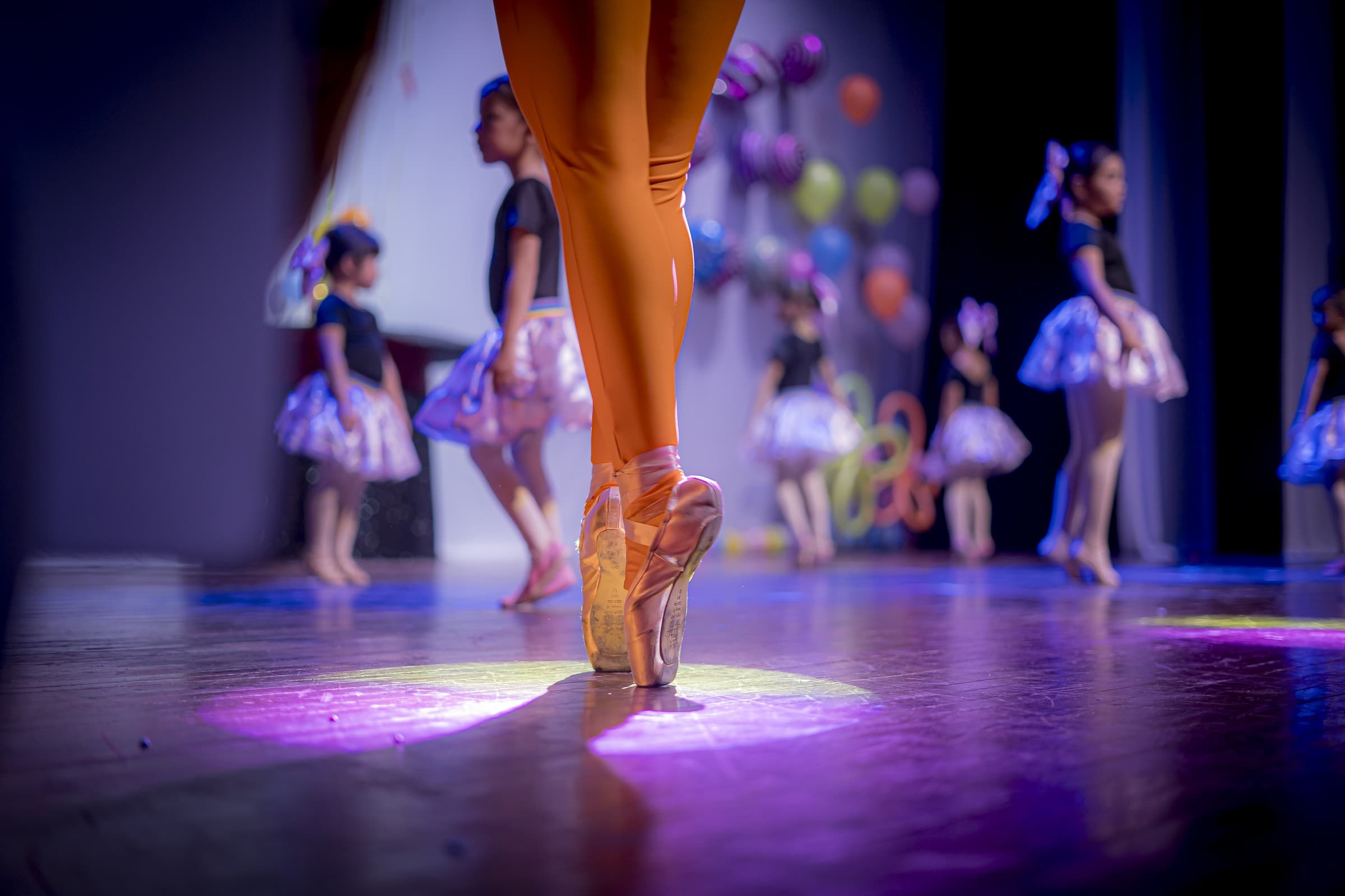 Bailarina en puntas bajo un spot de luz, con otras niñas bailarinas de fondo en el escenario